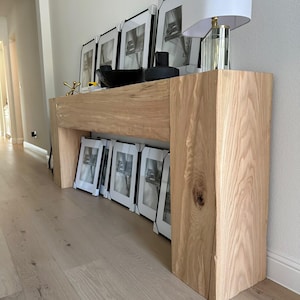 May include: A light wood console table with a natural finish. The table has a wide top and two legs. The table is decorated with framed black and white prints and a glass lamp with a white shade.