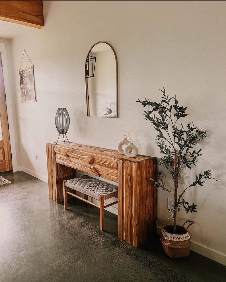 May include: A wooden console table with a natural wood finish and a woven bench. The table has a decorative arch mirror above it and a potted olive tree to the right.