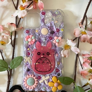 Könnte beinhalten: Eine transparente Handyhülle mit einem lila und weißen Design, das einen rosa Totoro-Charakter, Blumen und eine Schleife zeigt. Die Hülle hat einen schwarzen Gurt.