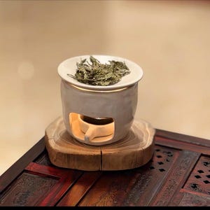 Op de afbeelding: Een witte keramische etherische olieverspreider met een ronde bovenkant met gedroogde groene bladeren. De diffuser staat op een houten voet en heeft een kleine opening aan de voorkant. De voet staat op een donkere houten tafel.