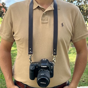May include: A brown leather camera strap with metal clasps, worn around the neck of a person wearing a beige polo shirt and blue jeans. A black Canon EOS 80D camera with a lens is attached to the strap.