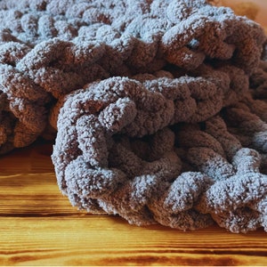 May include: A soft, grey, chunky knit blanket with a textured, woven pattern. The blanket is folded and draped over a wooden surface.