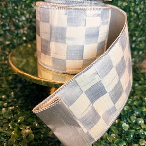 May include: A roll of decorative ribbon with a blue and white checkered pattern. The ribbon has a metallic silver edge and is displayed on a gold-colored stand against a backdrop of green foliage. The ribbon is approximately 2 inches wide.
