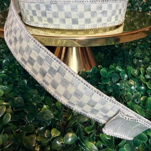 May include: A roll of decorative ribbon with a checkered pattern in shades of gray and cream. The ribbon has a metallic silver edge and is displayed on a gold-colored stand against a backdrop of green foliage. Perfect for crafts and gift wrapping.