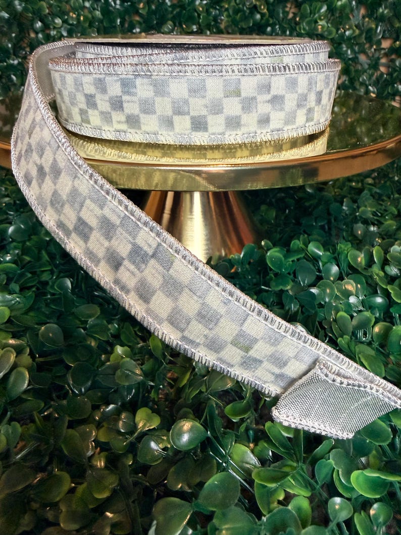 May include: A roll of decorative ribbon with a checkered pattern in shades of gray and cream. The ribbon has a metallic silver edge and is displayed on a gold-colored stand against a backdrop of green foliage. Perfect for crafts and gift wrapping.