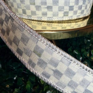 May include: A roll of decorative ribbon with a checkered pattern in shades of gray and cream. The ribbon has a metallic silver trim and is likely used for crafts, gift wrapping, or home decor projects.