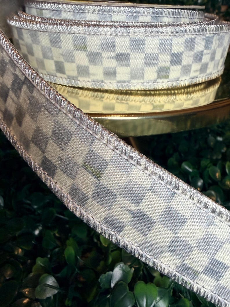 May include: A roll of decorative ribbon with a checkered pattern in shades of gray and cream. The ribbon has a metallic silver trim and is likely used for crafts, gift wrapping, or home decor projects.