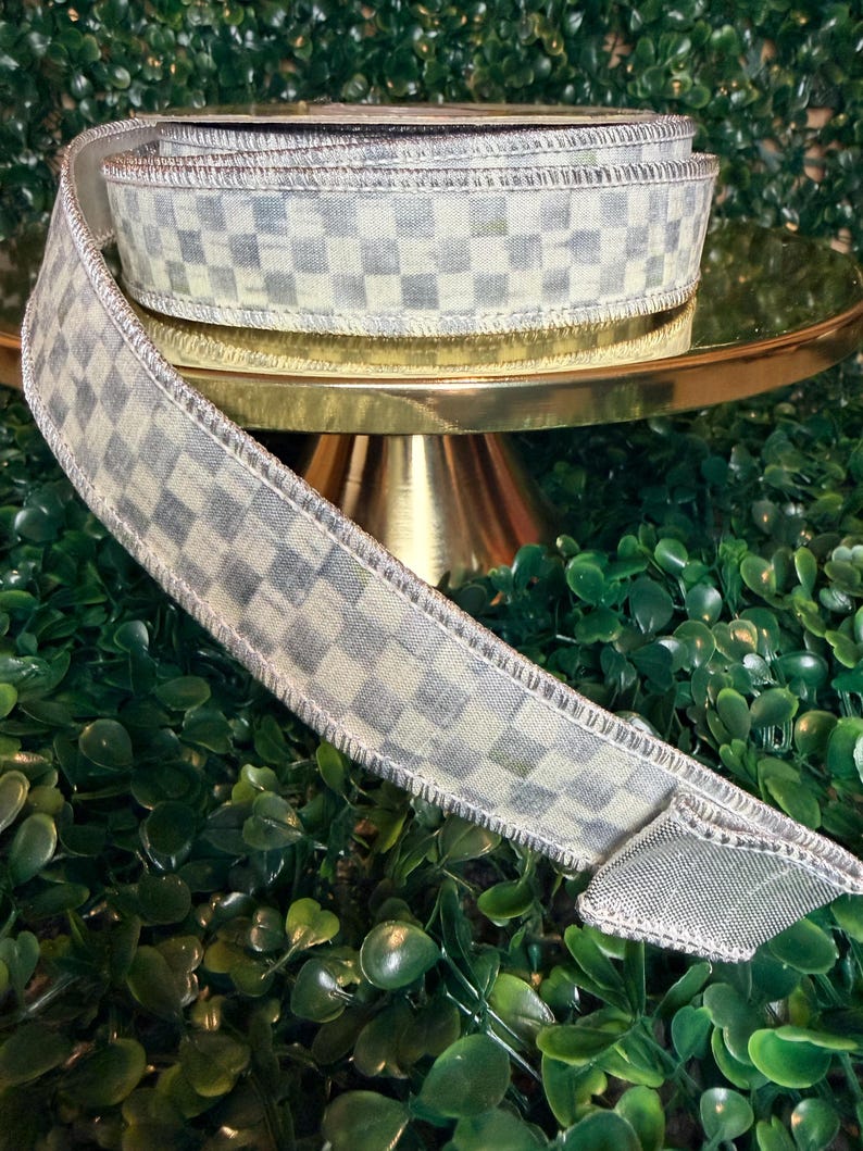 May include: A roll of decorative ribbon with a checkered pattern in shades of gray and cream. The ribbon is displayed on a gold-colored pedestal, with a green leafy background. The ribbon's edges are finished with a silver trim.