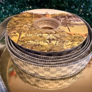 May include: A spool of decorative ribbon with a silver and white checkered pattern. The center of the spool features a landscape illustration with the text "MacKenzie-Childs". The ribbon is displayed on a gold surface.
