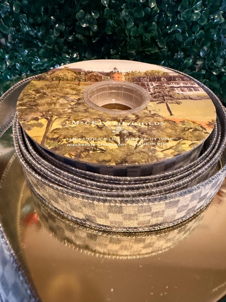May include: A spool of decorative ribbon with a silver and white checkered pattern. The center of the spool features a landscape illustration with the text "MacKenzie-Childs". The ribbon is displayed on a gold surface.