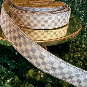 May include: A spool of decorative ribbon with a checkered pattern in shades of gray and white. The ribbon is displayed on a gold-colored stand, with a green background.