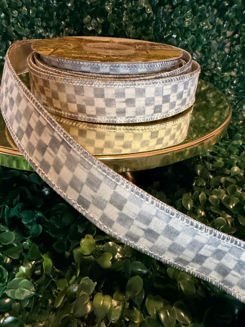 May include: A spool of decorative ribbon with a checkered pattern in shades of gray and white. The ribbon is displayed on a gold-colored stand, with a green background.