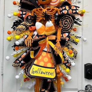 May include: A Halloween-themed wreath featuring a skeleton design with a carved pumpkin head. The wreath is adorned with candy corn, black and orange ribbons, and a sign that says "Halloween!"