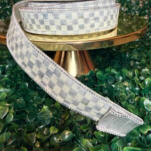 May include: A roll of decorative ribbon with a checkered pattern in shades of gray and white. The ribbon is displayed on a gold cake stand, with a green leafy background. The ribbon has a wired edge.