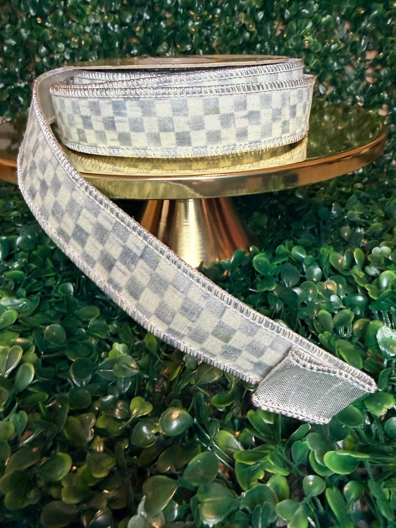 May include: A roll of decorative ribbon with a checkered pattern in shades of gray and white. The ribbon is displayed on a gold cake stand, with a green leafy background. The ribbon has a wired edge.