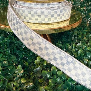 May include: A roll of decorative ribbon with a checkered pattern in shades of gray and cream. The ribbon has a metallic silver edge and is displayed on a gold-colored stand against a backdrop of green foliage. Ideal for crafts and gift wrapping.