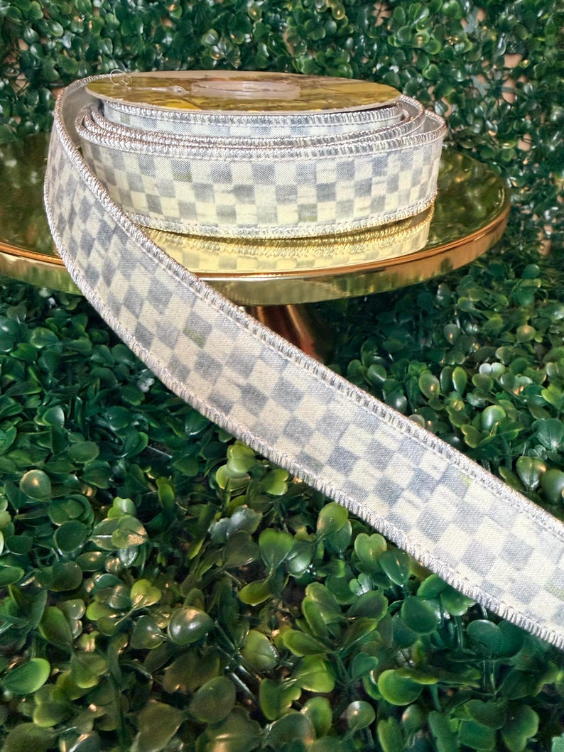 May include: A roll of decorative ribbon with a checkered pattern in shades of gray and cream. The ribbon has a metallic silver edge and is displayed on a gold-colored stand against a backdrop of green foliage. Ideal for crafts and gift wrapping.
