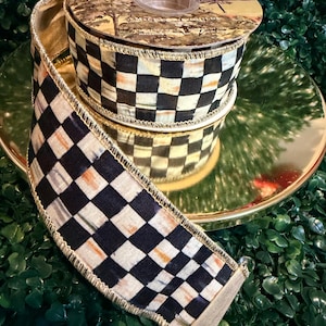 May include: A decorative ribbon spool with a black and white checkered pattern. The ribbon has a gold trim and a landscape design on the top. The spool is resting on a gold-colored plate.