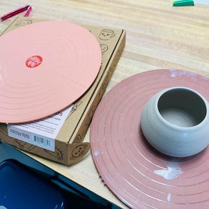 May include: A pottery wheel throwing kit with a pink splash pan and a gray ceramic bowl. The kit includes a pink splash pan with concentric circles and a red logo. A cardboard box is also visible.