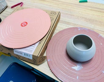 May include: A pottery wheel throwing kit with a pink splash pan and a gray ceramic bowl. The kit includes a pink splash pan with concentric circles and a red logo. A cardboard box is also visible.