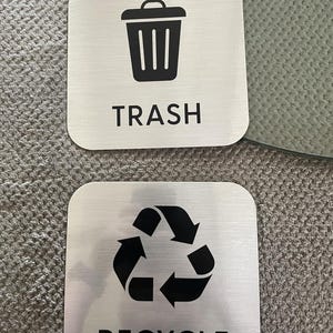 May include: Two square, brushed metal signs. One features a black trash can icon with the word "TRASH" below. The other has a black recycling symbol with the word "RECYCLE" below. Both signs have rounded corners.