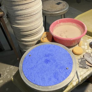 May include: A pottery studio scene featuring a blue pottery wheel, a stack of light-colored clay discs, and a pink basin filled with a light-colored liquid. The pottery wheel has a blue textured surface. Various tools and sponges are scattered around.