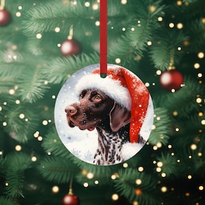 May include: A round Christmas ornament featuring a brown and white German Shorthaired Pointer dog wearing a red Santa hat. The dog is looking to the left and the background is a snowy scene.
