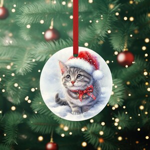 May include: A grey and white kitten wearing a red Santa hat with a red bow and bells around its neck. The kitten is sitting in the snow and looking to the right. The ornament is round and white with a snowy background.