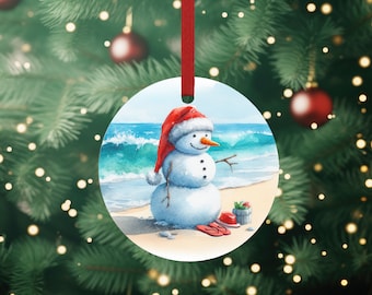 May include: A round Christmas ornament featuring a snowman wearing a red Santa hat and standing on a beach with blue water and waves. The snowman is holding a stick in one hand and has a carrot nose. There are red flip flops and a bucket with a green plant on the beach.