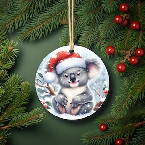 May include: A round Christmas ornament featuring a cartoon koala wearing a red Santa hat. The koala is sitting in a snowy forest setting.
