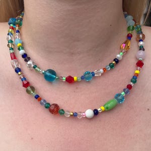 May include: A multi-layered necklace featuring colourful glass beads in shades of red, blue, green, yellow, and white. The necklace is made up of various sizes and shapes of beads, including round, oval, and square.