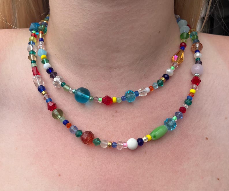 May include: A multi-layered necklace featuring colourful glass beads in shades of red, blue, green, yellow, and white. The necklace is made up of various sizes and shapes of beads, including round, oval, and square.