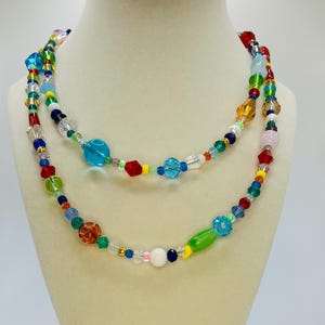 May include: A multi-strand necklace made of colourful glass beads. The beads are in various shapes and sizes, including round, oval, and square. The necklace is a vibrant mix of colours, including red, orange, yellow, green, blue, and purple.