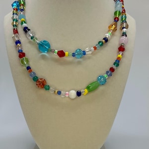 May include: A multi-strand necklace made of colourful glass beads. The necklace features a variety of shapes and sizes of beads, including round, oval, and square beads. The beads are strung on a clear cord and are arranged in a random pattern. The necklace is a vibrant and eye-catching piece of jewellery.