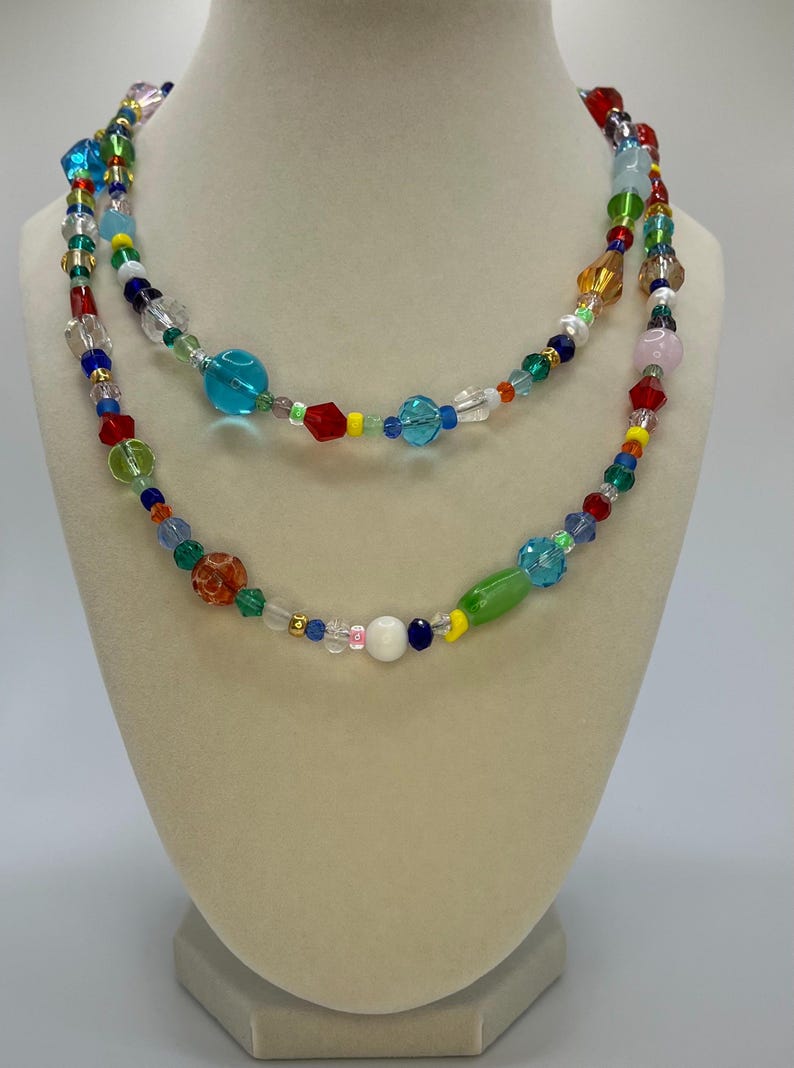 May include: A multi-strand necklace made of colourful glass beads. The necklace features a variety of shapes and sizes of beads, including round, oval, and square beads. The beads are strung on a clear cord and are arranged in a random pattern. The necklace is a vibrant and eye-catching piece of jewellery.