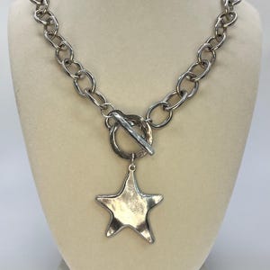 May include: Silver-toned necklace featuring a large star pendant. The necklace has a chunky chain design with a toggle clasp. The star pendant is a five-pointed shape, adding a bold statement to the jewelry piece. The necklace is displayed on a mannequin.