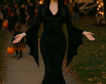 Puede incluir: Un vestido negro de terciopelo con mangas de murciélago. El vestido tiene un cuello en V y una silueta de sirena. La mujer lleva el vestido en una acera con un fondo borroso de gente y decoraciones de Halloween.