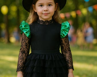 May include: A young girl wearing a black witch costume with a black hat, a black dress with lace sleeves, and green satin accents on the shoulders. The dress has a tiered skirt and a black ribbon belt. The background is a blurred outdoor scene.
