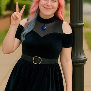 May include: A woman wearing a black velvet dress with a sheer mesh neckline and a gold belt. She is wearing a blue pendant necklace and has pink and blue hair.