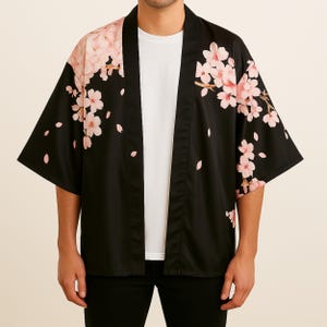 Puede incluir: Una kimono negra con estampado de flores de cerezo rosas. La kimono tiene mangas cortas y un estilo japonés tradicional.