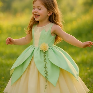 Puede incluir: Una niña pequeña con un vestido de princesa verde y amarillo con un adorno de flor y vid. El vestido tiene una falda de tul y un corpiño con un diseño de nenúfar.