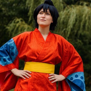 Könnte beinhalten: Eine Person trägt einen leuchtend roten Kimono mit einem gelben Obi-Gürtel. Der Kimono hat blaue Ärmel mit einem kreisförmigen Muster. Die Person trägt auch eine schwarze Perücke. Der Hintergrund ist ein verschwommenes Grün.