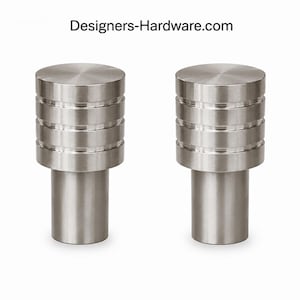 May include: Two brushed nickel cabinet knobs. Each knob has a cylindrical base and a stacked, tiered design on the upper portion. The knobs are made of metal and have a modern, minimalist aesthetic. The website Designers-Hardware.com is visible at the top.