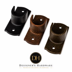 May include: Three metal curtain rod brackets featuring a U-shaped cradle and a rectangular mounting plate with two screw holes. The set includes one matte black, one rustic copper, and one antique bronze finish. The logo DH Designer's Hardware is below.