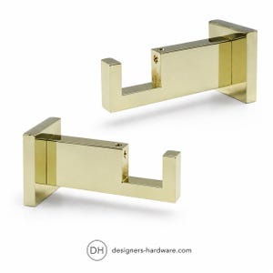 May include: Two gold-toned metal wall hooks with a square base and a rectangular arm featuring a small upward-facing hook at the end. Each piece has a visible screw hole on the top surface. The text DH designers-hardware.com is at the bottom.