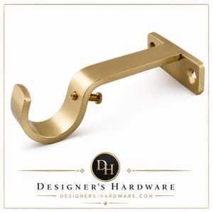May include: A brushed brass curtain rod bracket featuring a curved support arm, a circular cradle for the rod, and a small adjustment screw on the side. The mounting plate includes two pre-drilled holes for installation against a flat surface.