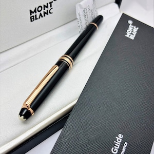 Può includere: Una penna Montblanc nera con dettagli color oro è nella sua scatola di presentazione. La penna presenta una clip e finiture dorate, con il logo Montblanc sulla punta. La scatola è bianca con il logo Montblanc stampato in nero.