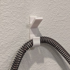May include: White plastic hook mounted on a textured wall with a coiled black hose hanging from it.