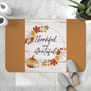 Op de afbeelding: Een bruine en oranje deurmat met een witte achtergrond met een krans van herfstbladeren en de tekst "Thankful and Grateful".