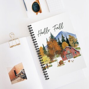 May include: A spiral bound notebook with a watercolor illustration of a red barn, trees, and mountains. The text "Hello Fall" is written in black cursive at the top of the notebook.